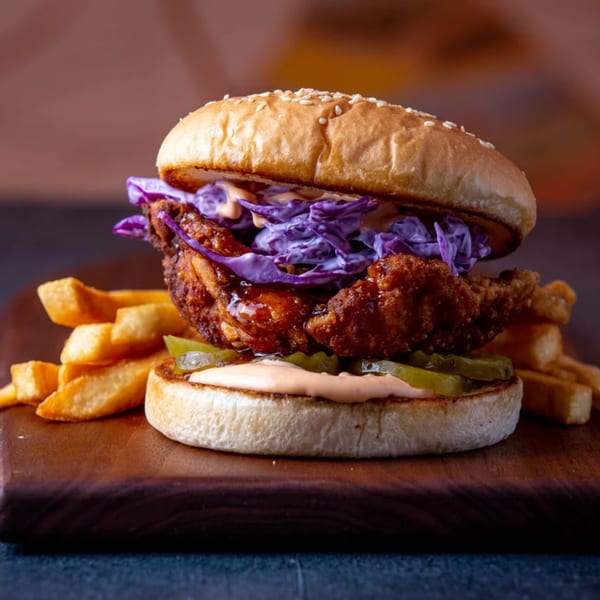 Dusty's Hideaway fried chicken sandwich with purple slaw, pickles, sauce, and fries on a wooden board.