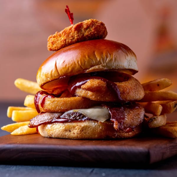 A Dusty's Hideaway burger with bacon, onion rings, cheese, BBQ sauce, and a chicken nugget on top, served with fries.
